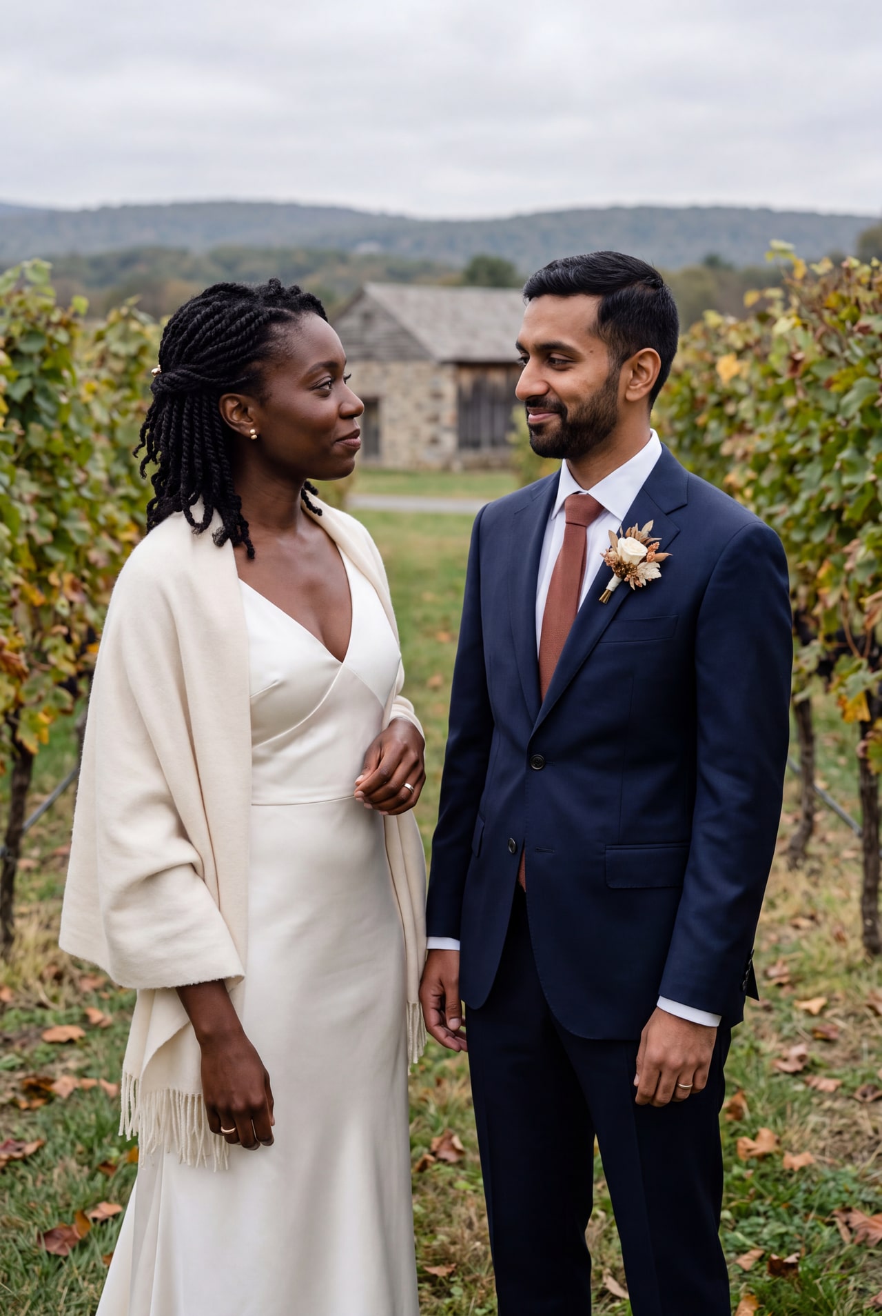 A bride in an ivory dress and wool wrap beside a navy-suited groom in an autumn vineyard