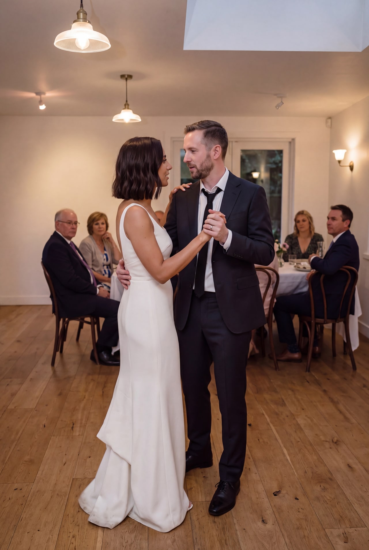 A bride and groom dancing while guests sit around a softly lit reception room