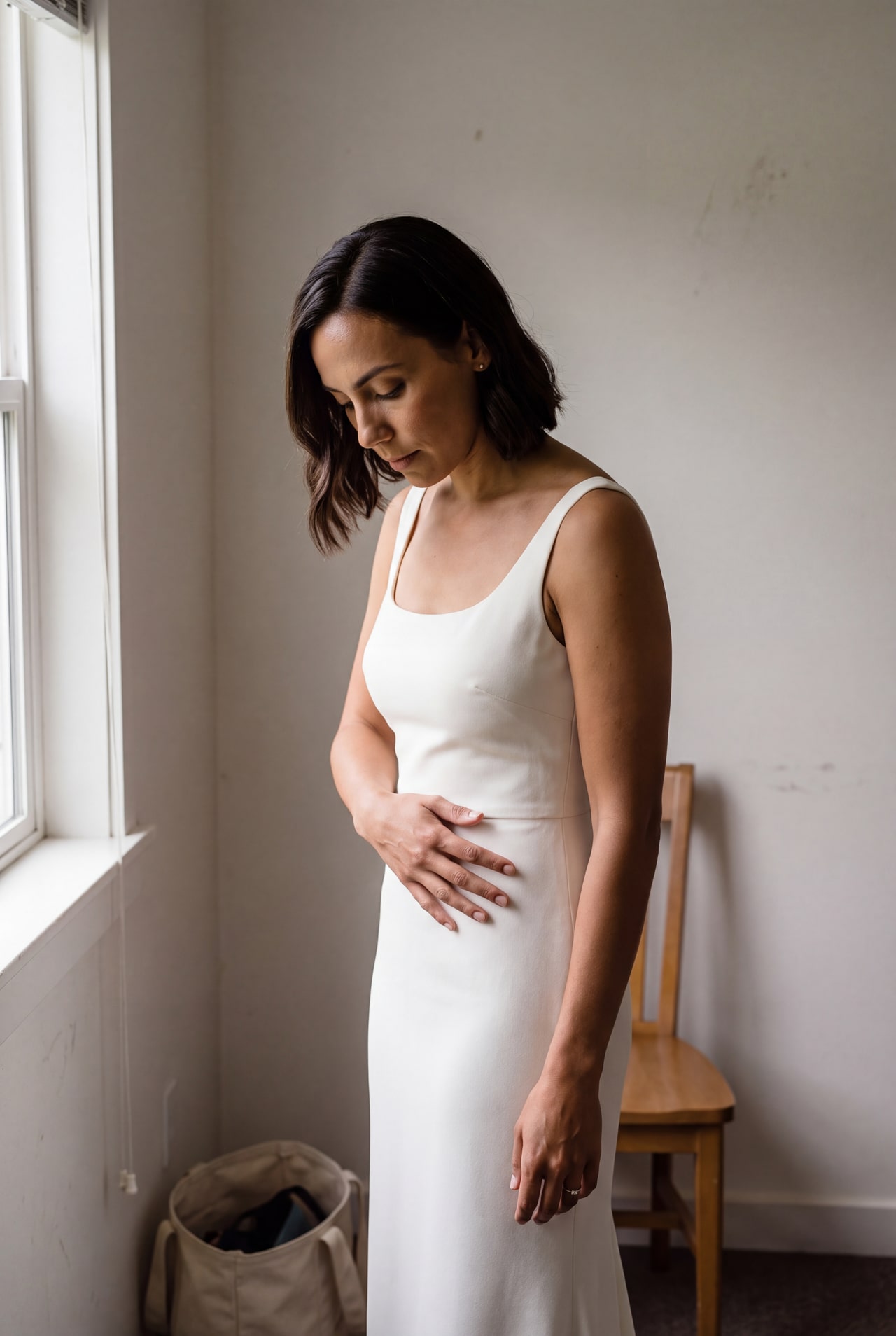 A bride pausing beside a plain window in her ivory gown