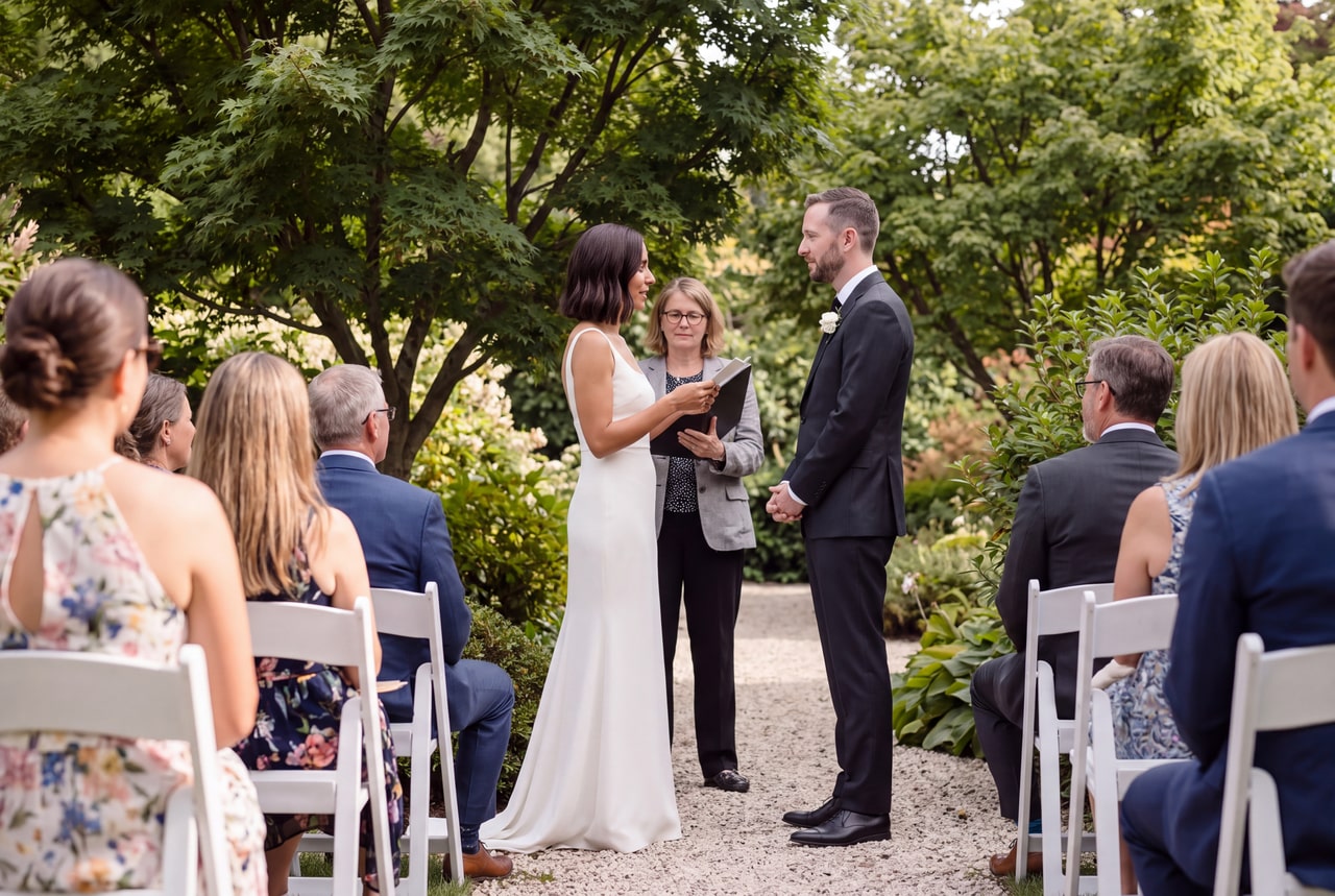 A bride reading from a small vow card beside the groom in a garden ceremony