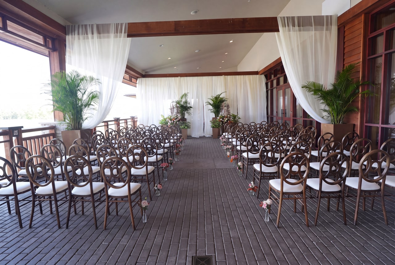 Covered terrace ceremony with palms, bronze chairs and an aisle beside dark timber windows at Legends on the Niagara.