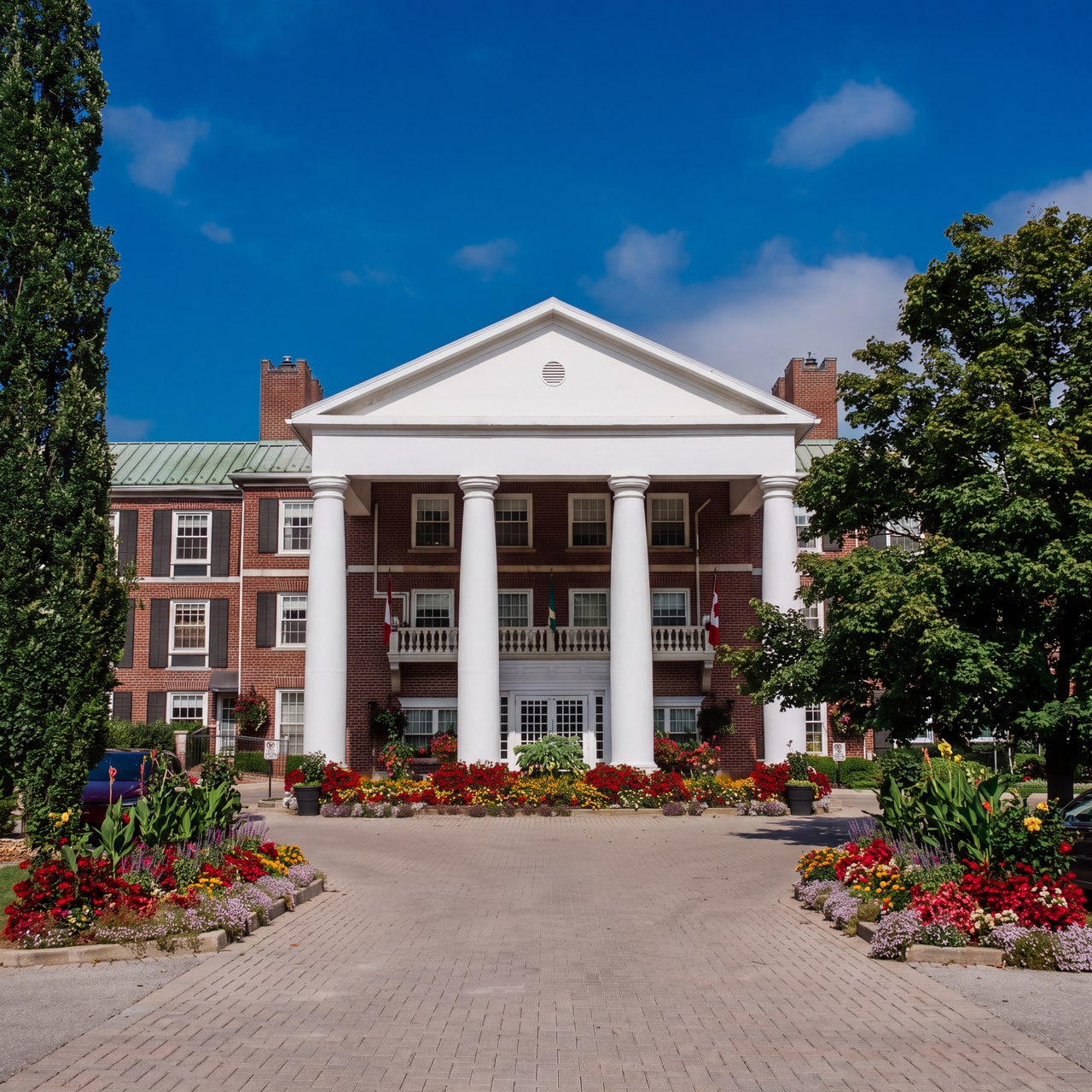 White columns and a balustraded balcony above the red-brick entrance and flower beds at Queen’s Landing.