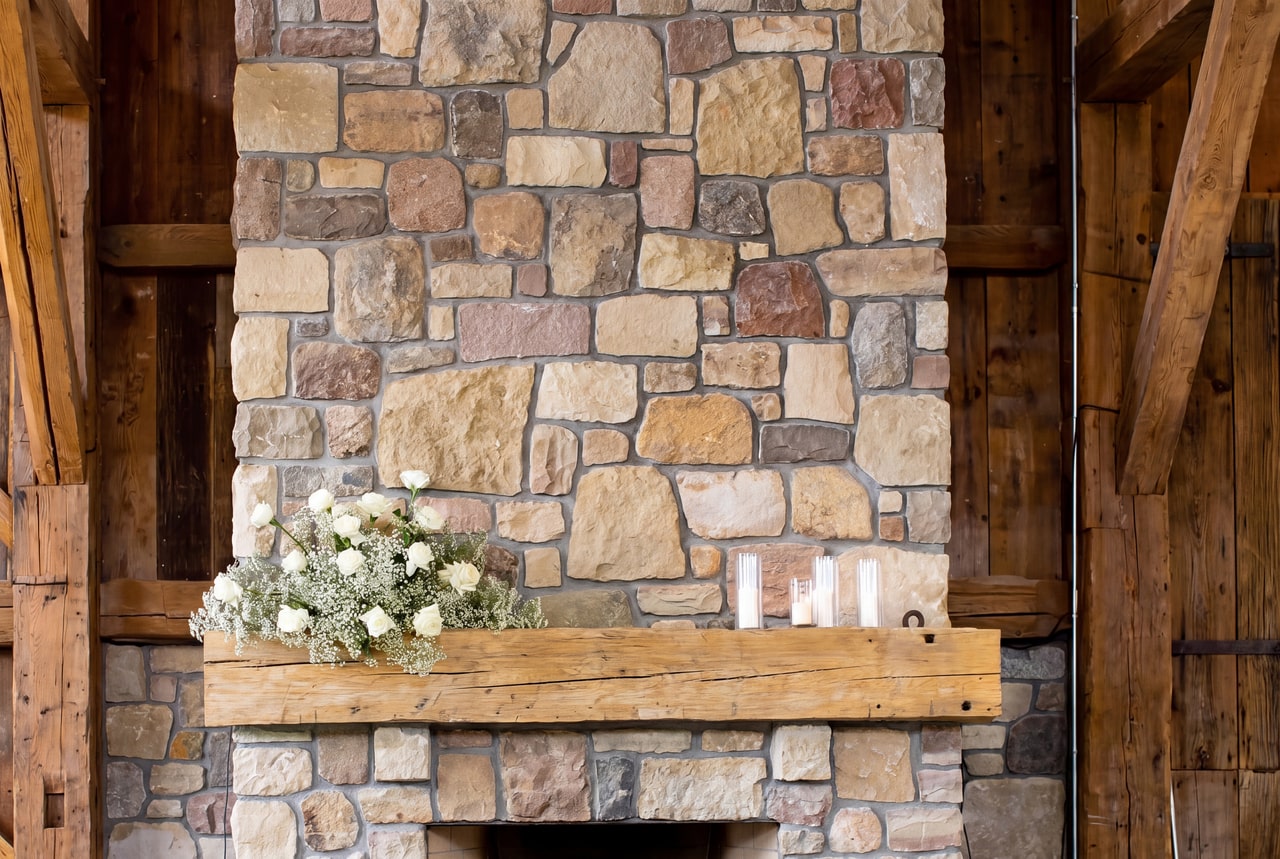 Rough timber mantel, stone chimney breast and white flowers inside Legacy Hall at Vieni Estates.
