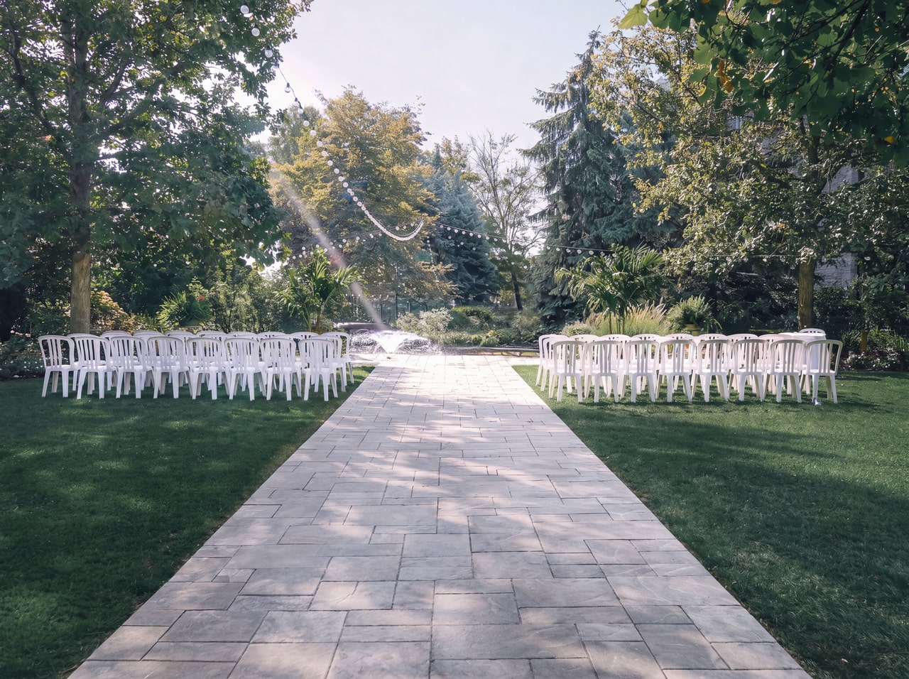 White ceremony chairs beside a pale paved aisle, mature trees and a low garden fountain at White Oaks.