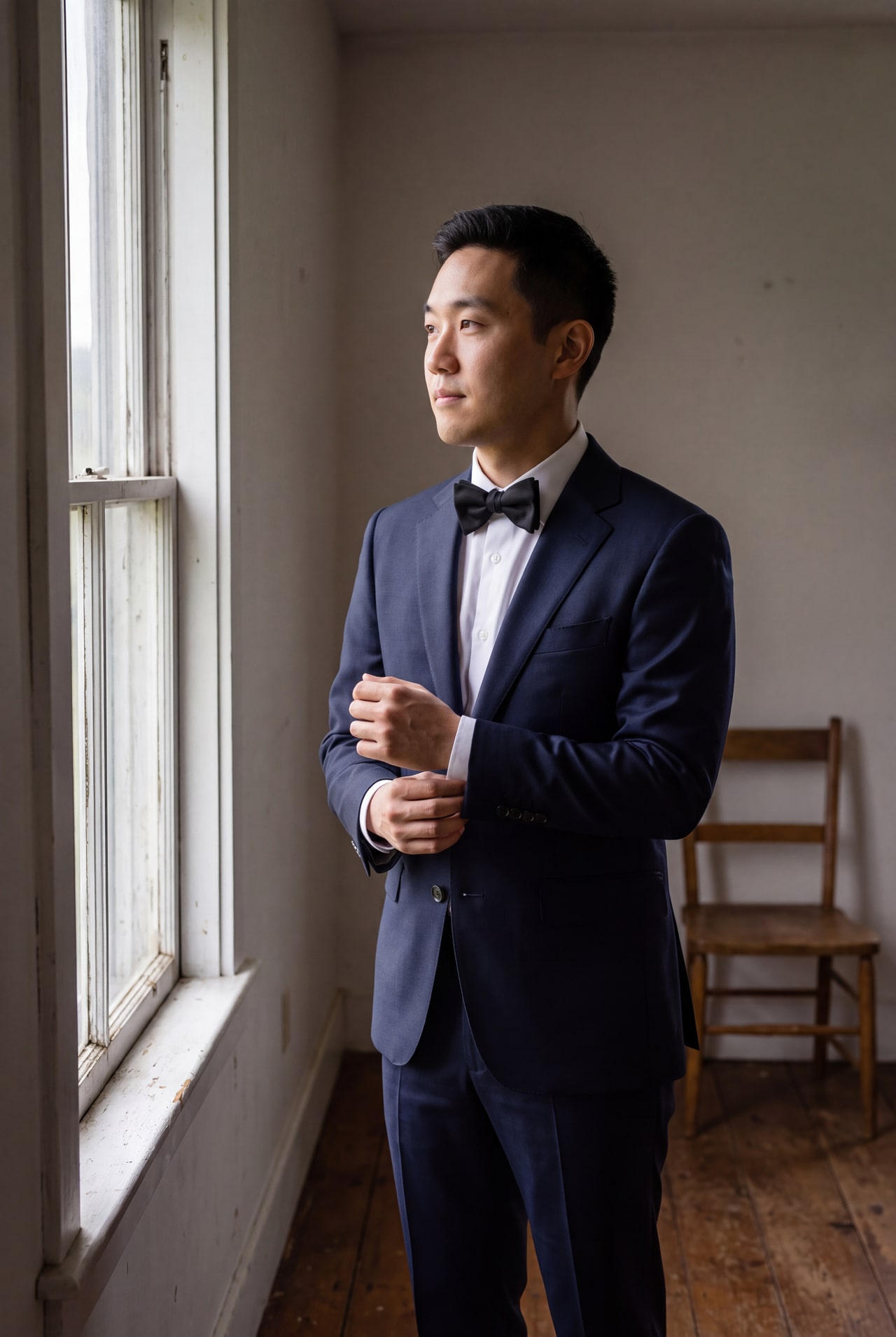 A groom adjusting his cuff beside a sash window in a simple preparation room