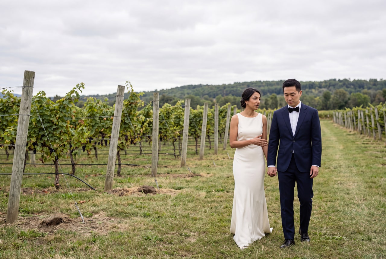 A bride and groom standing between working vineyard rows beneath an overcast sky